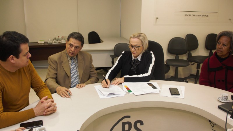 Quatro pessoas (dois homens e duas mulheres) sentadas atrás de uma mesa. Secretária Arita Assina documento. Um participantes da reunião fala enquanto os outros o escutam.