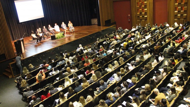 Foto tirada do alto do auditório mostra algumas pessoas sentadas em cadeiras no palco e a plateia assistindo.    