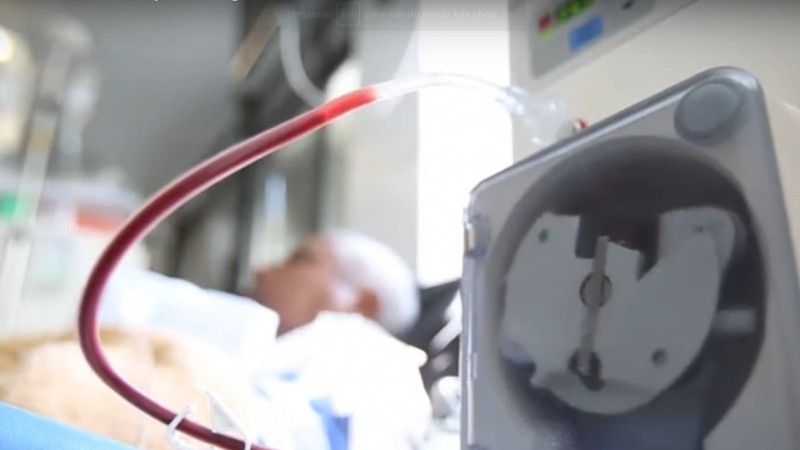 A imagem mostra, em primeiro plano, um aparelho fazendo a transfusão de sangue para um homem que está deitado na maca.   