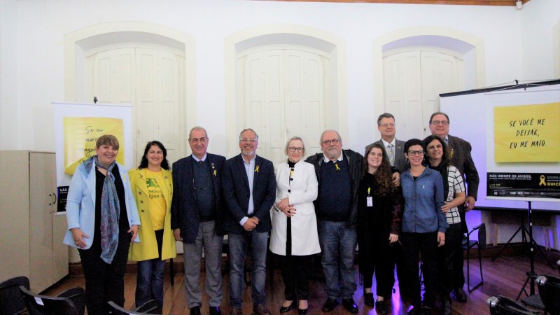Secretária Arita (no centro) e representantes das entidades parceiras no lançamento da campanha. Todos estão de pé e sorriem. Atrás deles, tem dois banners. 