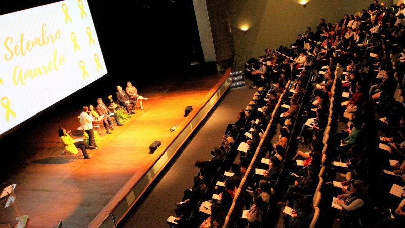 Foto tirada do alto do auditório mostra a plateia e o palco, com os participantes do evento. Atrás dos participantes, tem um telão com o mensagem "Setembro Amarelo" e laços amarelos.