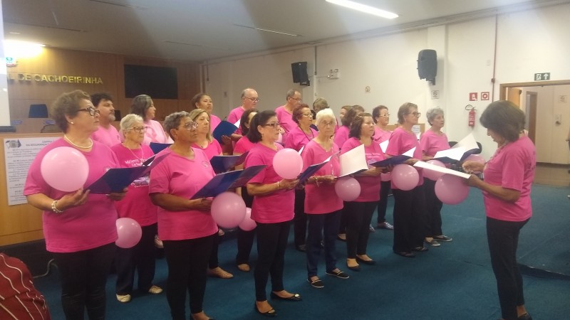 Várias pessoas de pé, com camiseta cor-de-rosa, cantando. cada um segura uma pasta aberta na mão (onde tem as letras das músicas) e um balão.