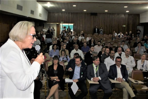 Secretária Arita fala, segurando o microfone. Sentada, a plateia assiste.