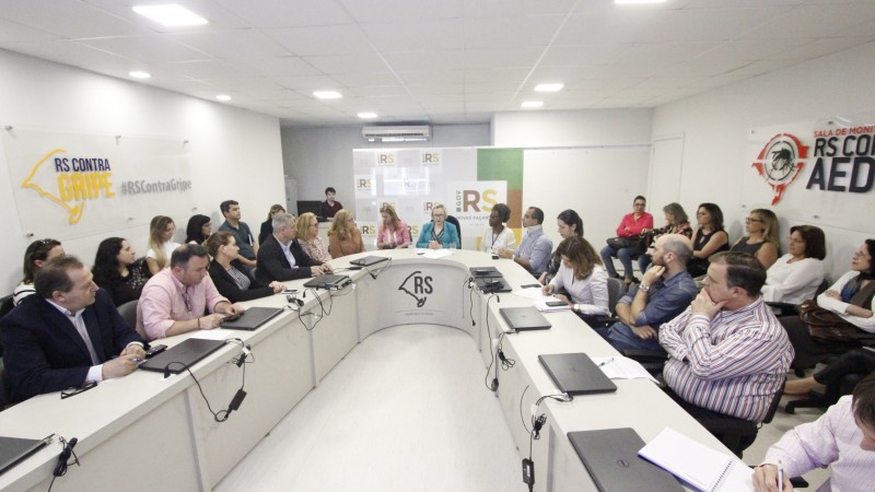 A imagem mostra a reunião do comitê, com várias pessoas sentadas atrás em uma mesa em formato de U. 