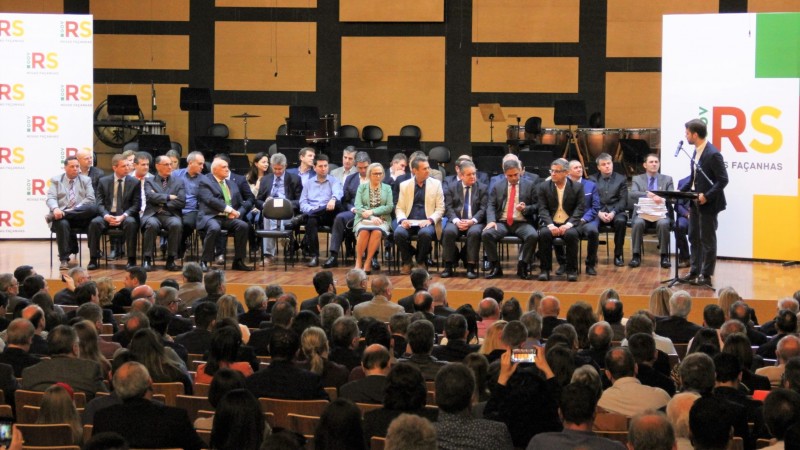 Várias pessoas sentadas no palco, entre elas a Secretária Arita, e na plateia observam o homem que fala de pé, atrás de um microfone. 